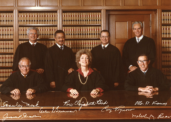 Autographed group portrait of Chief Justice Rose Bird with Associate Justices Otto Kaus, Malcom Lucas, Cruz Reynoso, Allen Broussard, Joseph Grodin, and Stanley Mosk. Chief Justice Bird is seated in the center of the group. The photograph was taken in the chief justice’s chambers, which are lined with built-in bookcases.