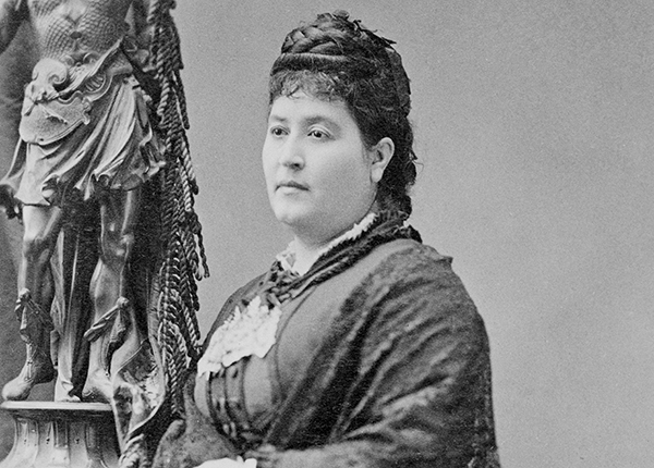 Cabinet card portrait of María Amparo Ruiz de Burton in a fancy Victorian gown, holding a shawl. Her hair is elaborately coiled on top of her head and her expression is somber. She is posing in a studio, with a classical statute of a male figure on a pedestal in the background.