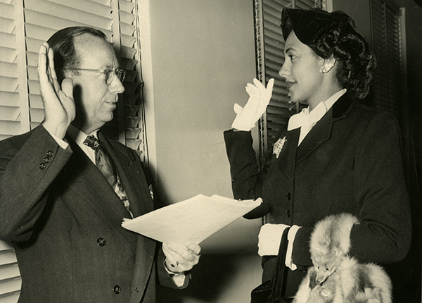 Photograph of Vaino Spencer being sworn in by an unidentified judge. Both Spencer and the judge are holding up their right hands. Spencer is stylishly dressed, wearing a black suit and hat, white gloves, and a fur draped over her left arm.