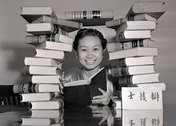 Photograph of Chiyoko Sakamoto seated at a table, her head framed by an arch made up of piles of law books. Sakamoto is holding an open book in her hands and smiling happily. A card with Japanese characters stands on the table.